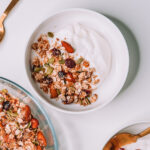 Plain yogurt with granola in a white bowl on a white table with two other bowls