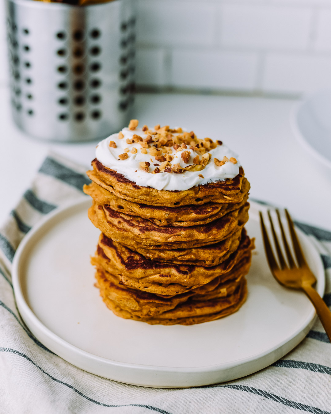 Stack of oatmeal pancakes topped with vanilla skyr and chopped peanuts on a white plate sitting on top of a kitchen towel on a white table. 