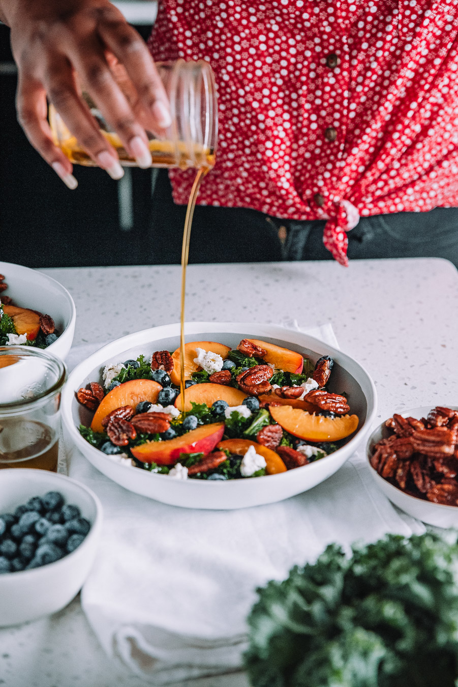 Woman pour dressing over a bowl filled with kale, peach, blueberry salad. 