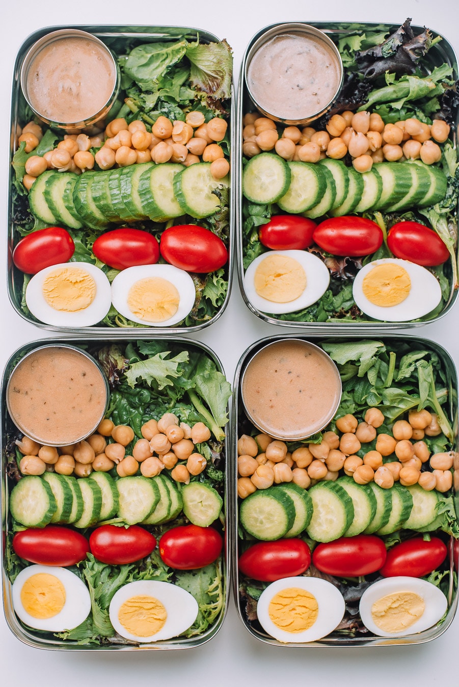 4 Bento boxes filled with mixed greems, boiled egg, grape tomatoes, mini cucumbers, chickpeas, and Drew's Organics Tahini Goddess Dressing and Marinade