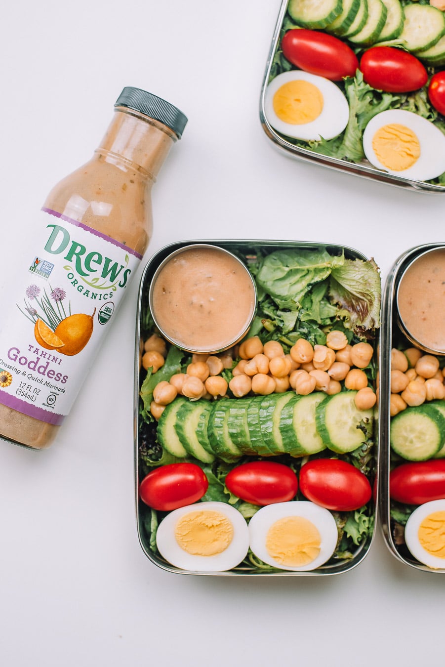 3 Bento boxes filled with mixed greens, boiled egg, grape tomatoes, mini cucumbers, chickpeas, and Drew's Organics Tahini Goddess Dressing and Marinade with bottle of dressing