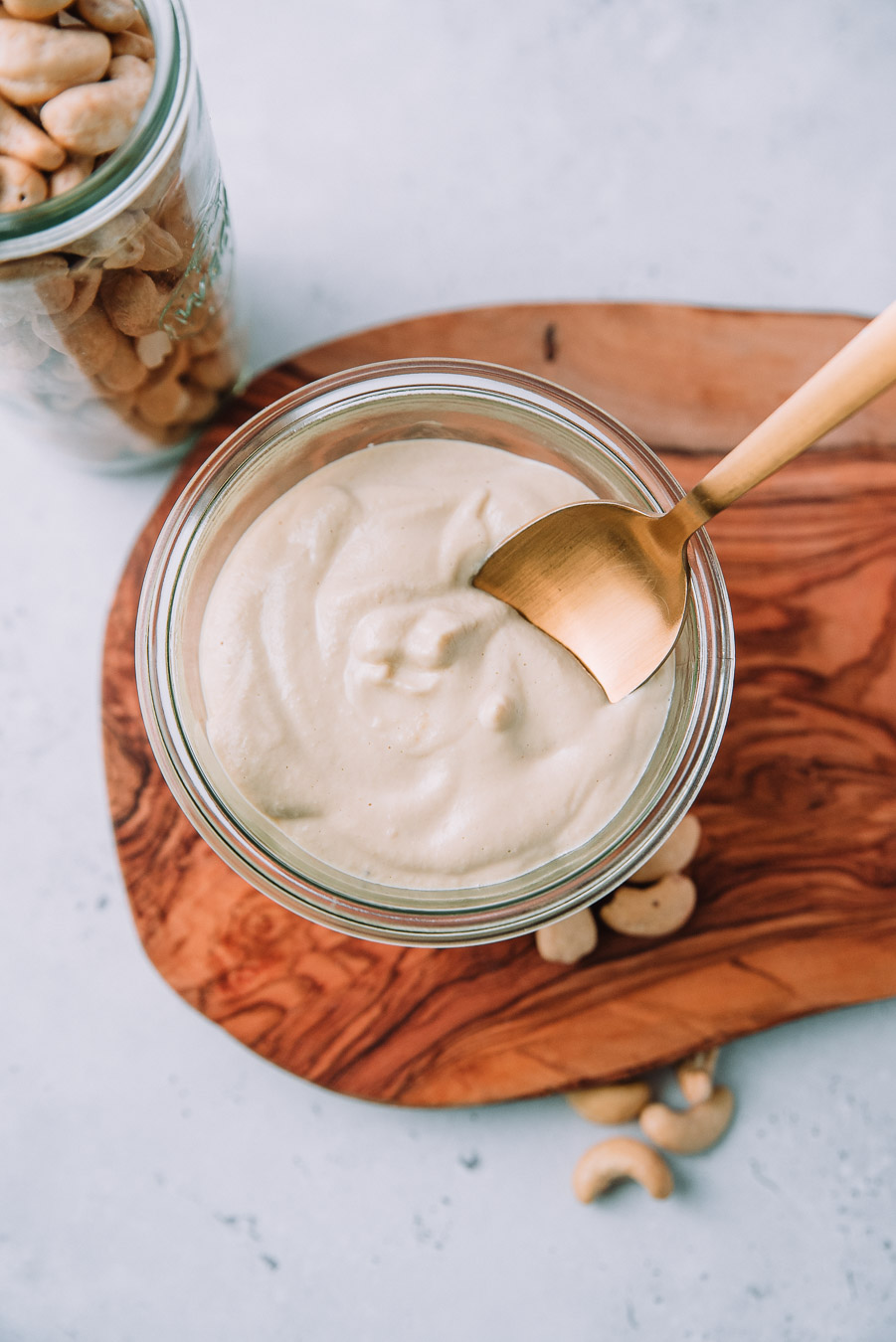 Cashew Alfredo Sauce in a jar with a gold spoon next to a jar of cashews, lemon, and a bulb of garlic