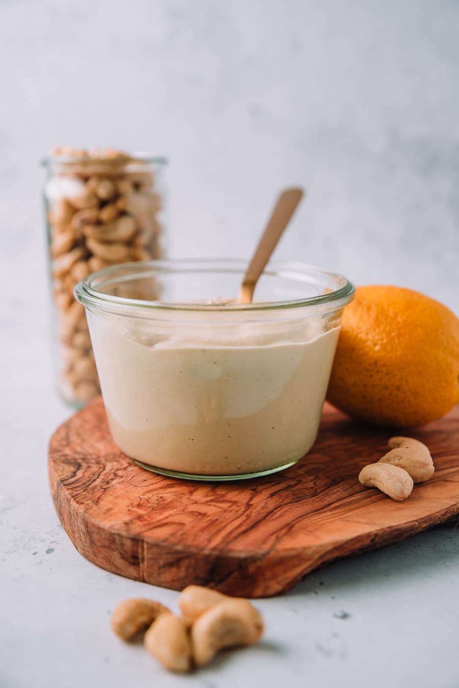 Cashew Alfredo Sauce in a jar with a gold spoon next to a jar of cashews, lemon, and a bulb of garlic