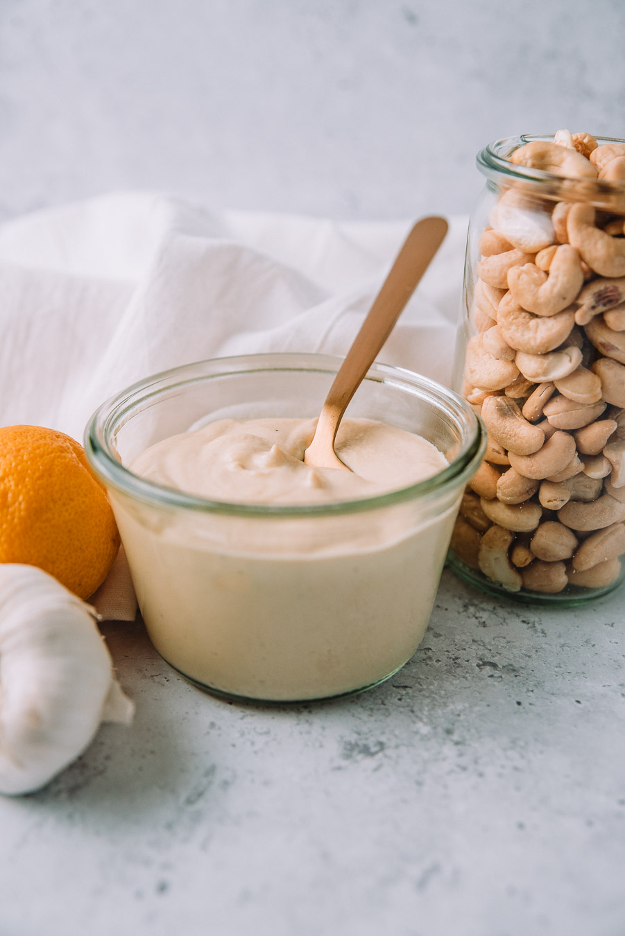 Cashew Alfredo Sauce in a jar with a gold spoon next to a jar of cashews, lemon, and a bulb of garlic