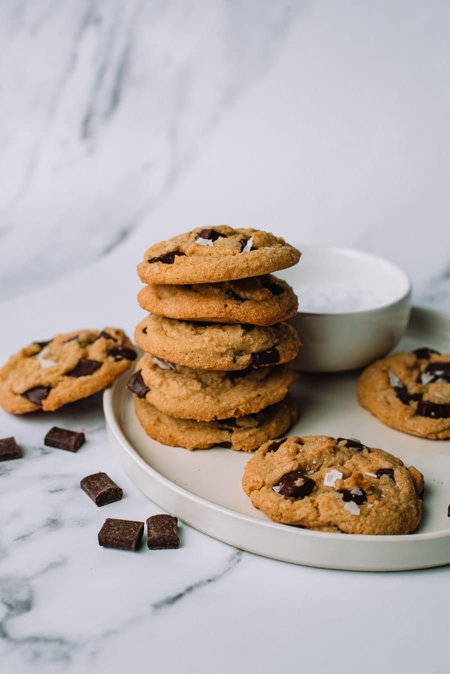 Olive Oil Chocolate Chip Cookies stacked on top of each other on a white plate with extra chocolate morsels.