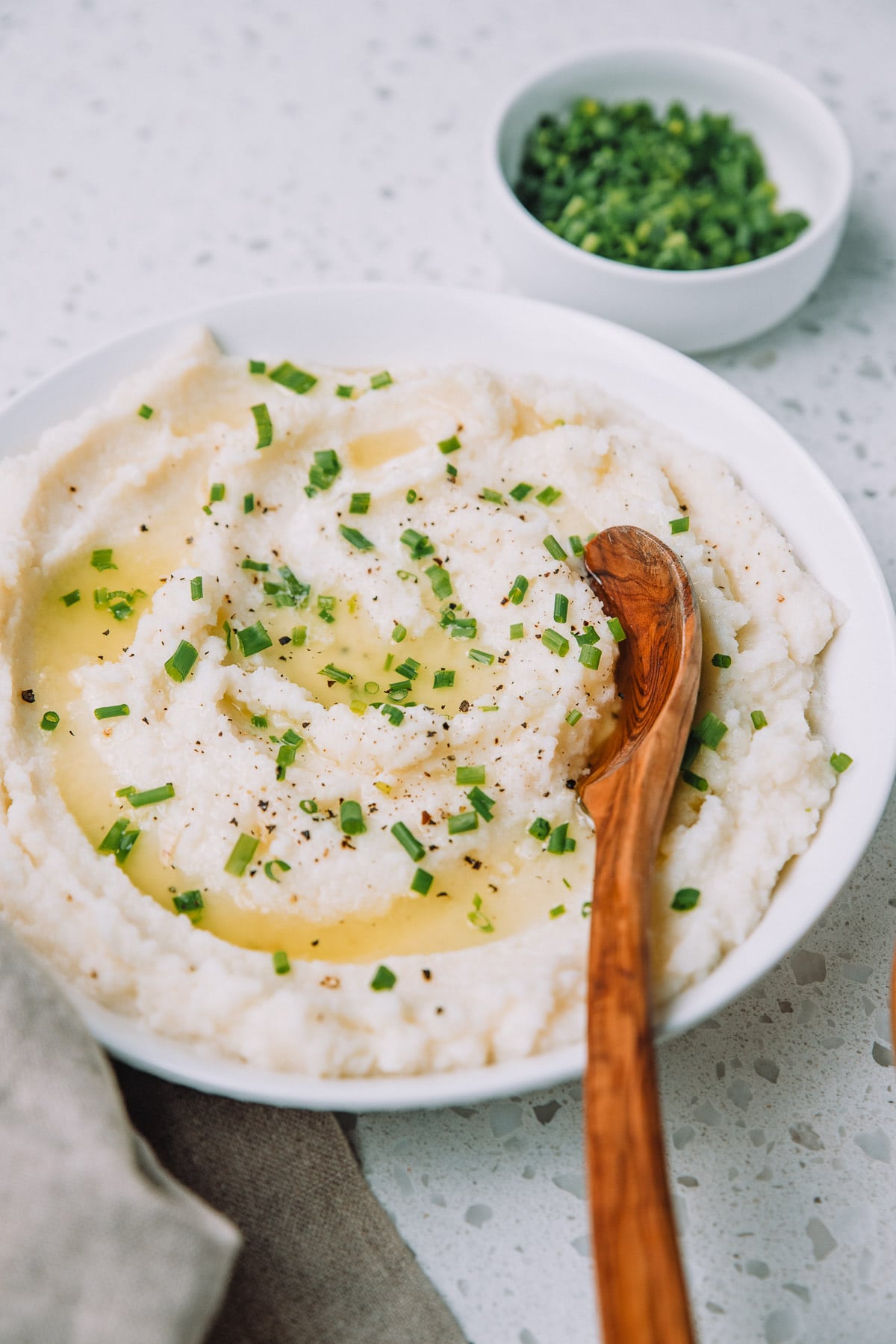 White bowl of roasted garlic mashed cauliflower topped with butter and chives with a wood spoon.