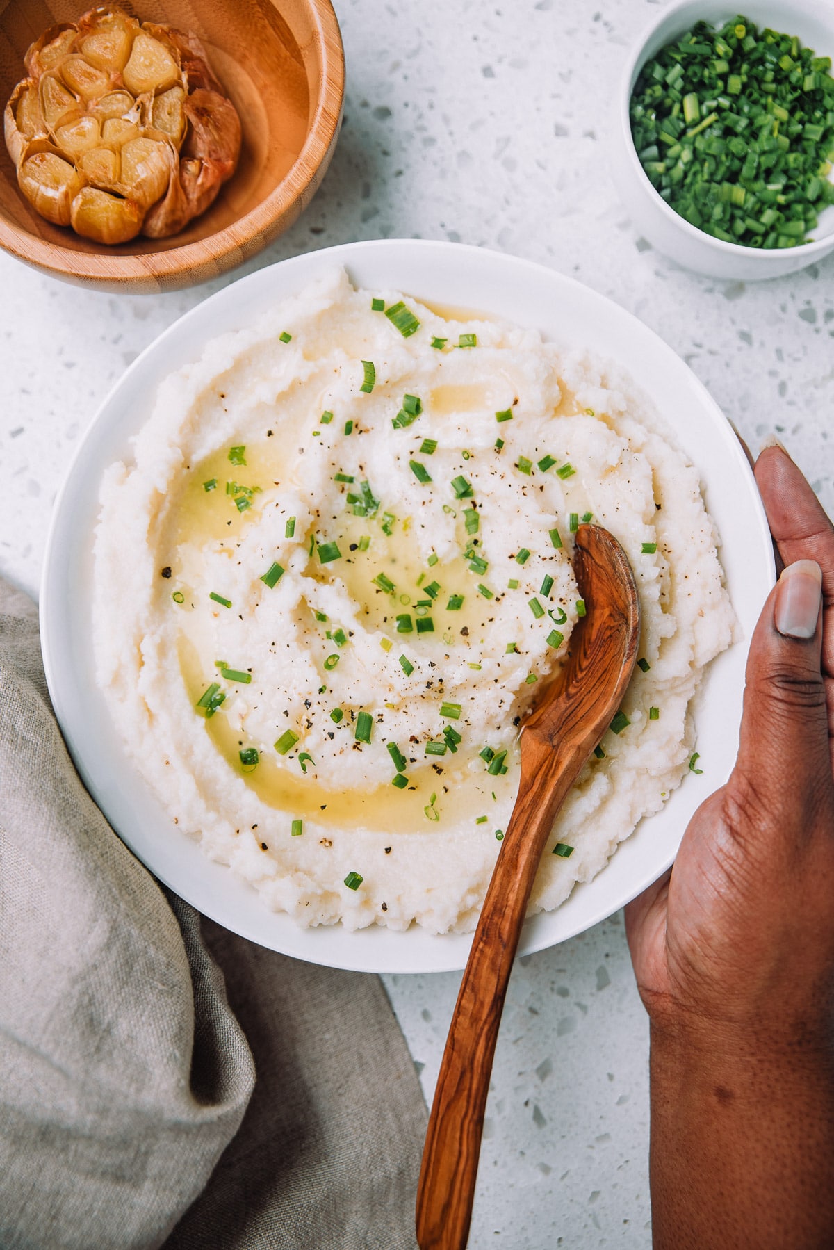 White bowl of roasted garlic mashed cauliflower topped with butter and chives on a table.